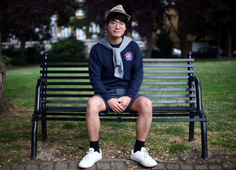 سيمون تشينج يتظاهر لالتقاط صورة.  يجلس على مقعد في حديقة في لندن.  كان يرتدي شورتاً أزرق وقميصاً من النوع الثقيل وقبعة.